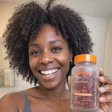 A smiling woman with curly hair holds up a bottle of Bloom & Bond pumpkin seed oil supplements.