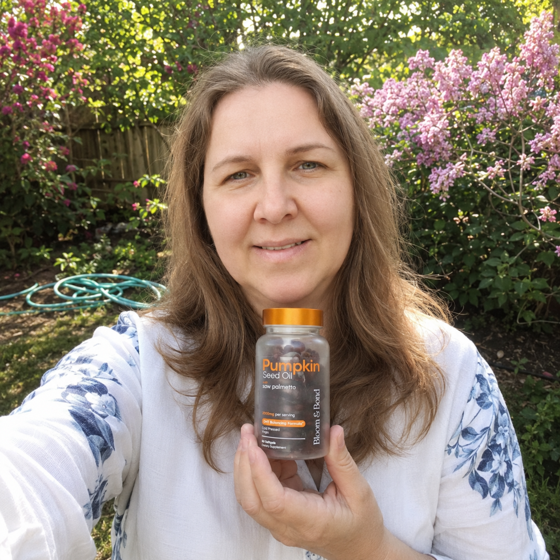 A smiling woman holds a bottle of Pumpkin Seed Oil supplements while standing in a garden.