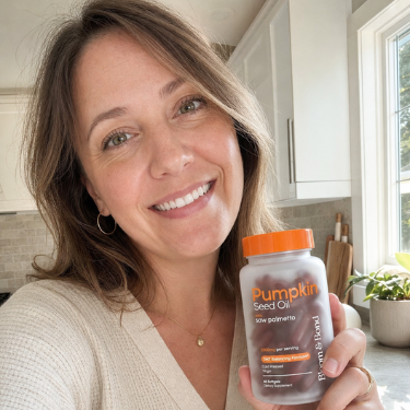 A smiling woman in a kitchen holds up a bottle of Pumpkin Seed Oil supplements.