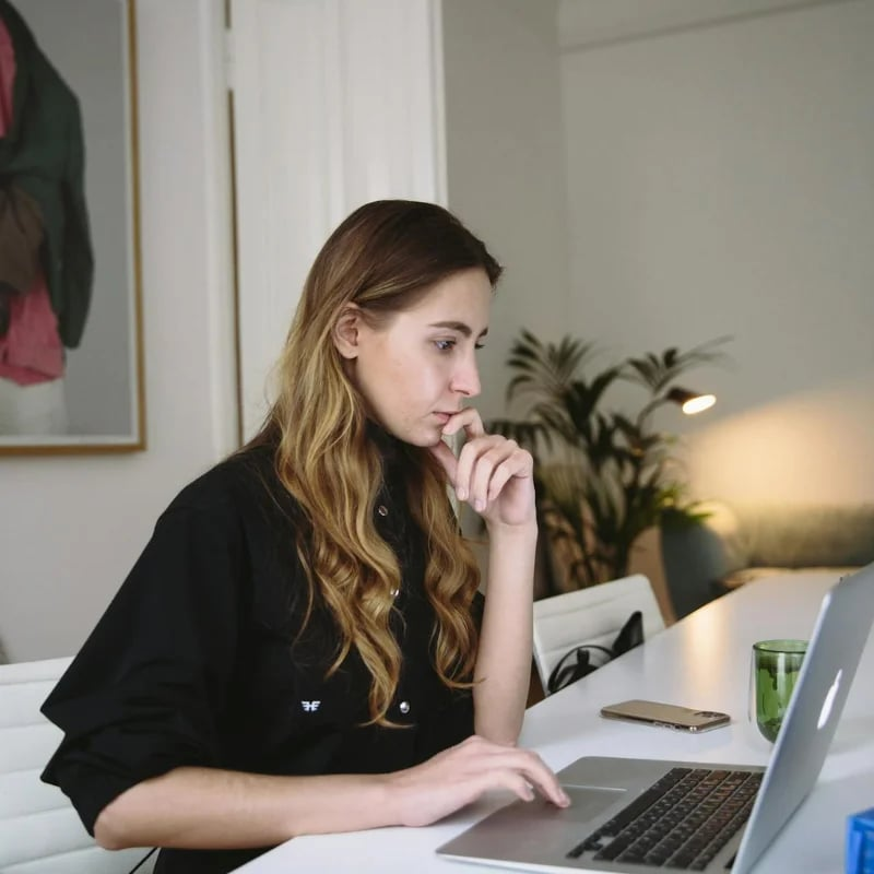 A woman in a black shirt sits at a white desk, looking thoughtfully at her laptop computer.