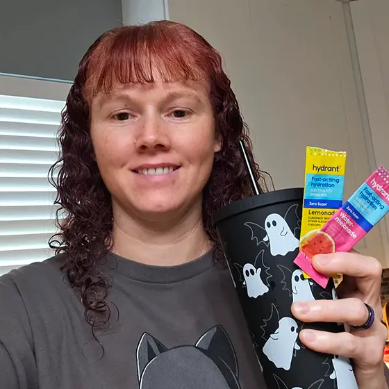 A smiling woman holds a black tumbler with ghosts and two packets of Hydrant drink mix.