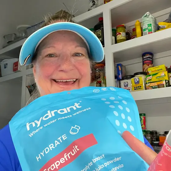 A smiling woman in a visor holds a large bag of Hydrant Grapefruit electrolyte powder in a pantry.