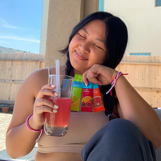 A smiling woman with braided hair holds a glass of pink drink and several drink mix packets.