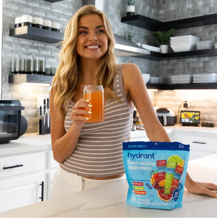 A smiling woman in a kitchen holds a glass of orange drink next to a bag of Hydrant.