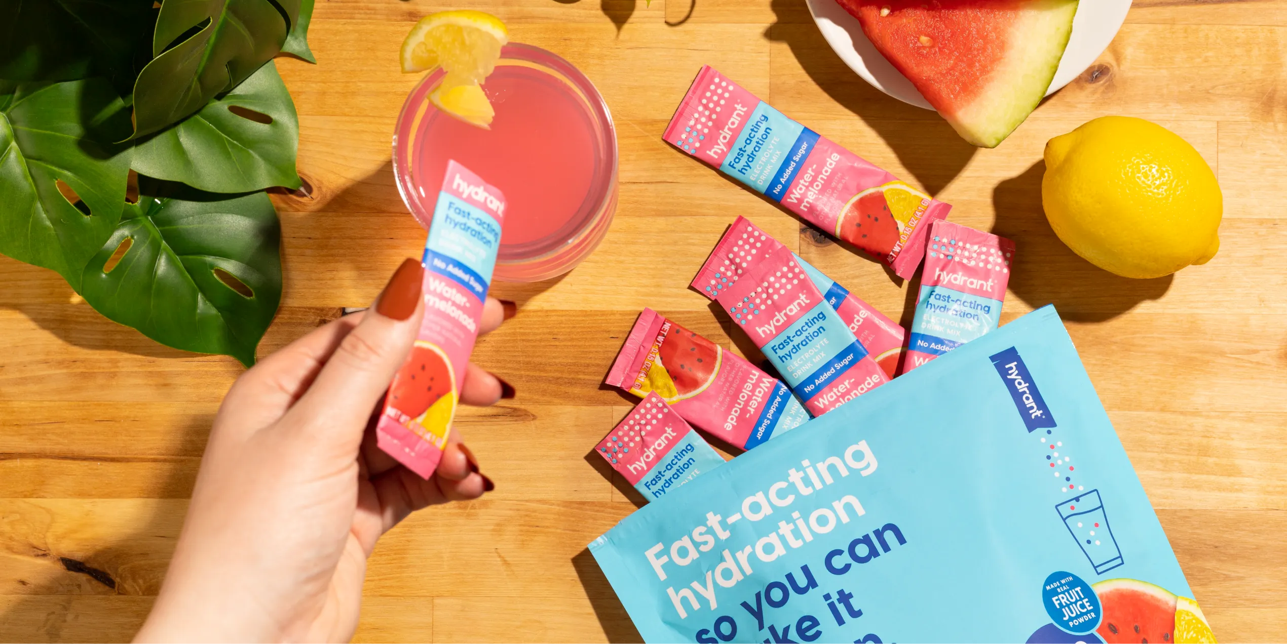 A hand holding a Hydrant Water-melonade packet over a table with a pink drink and fresh fruit.