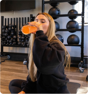 Person drinking orange beverage in a gym with exercise balls in the background.