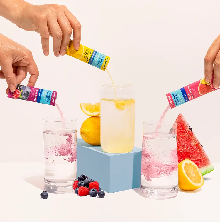 Hands pour flavored drink mix powders from packets into three glasses of water with corresponding fruits nearby.