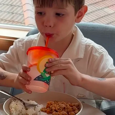 A young boy in a white collared shirt drinks from a colorful cup with a red straw.