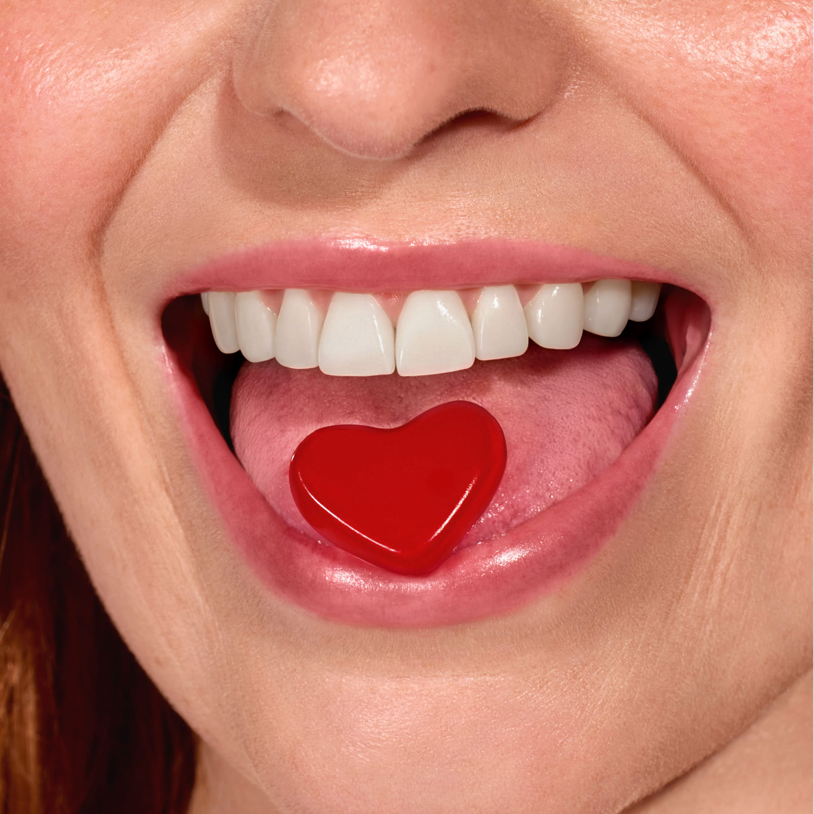 A close-up of a smiling woman's mouth with a red, heart-shaped candy on her tongue.