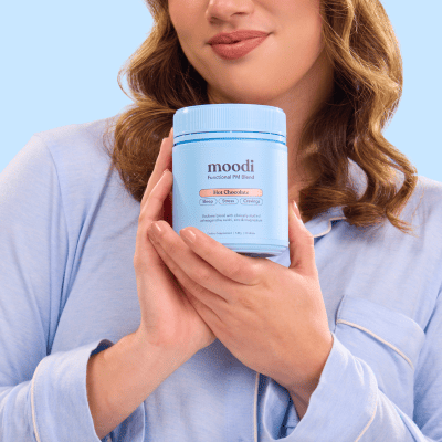 A woman in blue pajamas holds a light blue jar of Moodi functional PM blend hot chocolate.