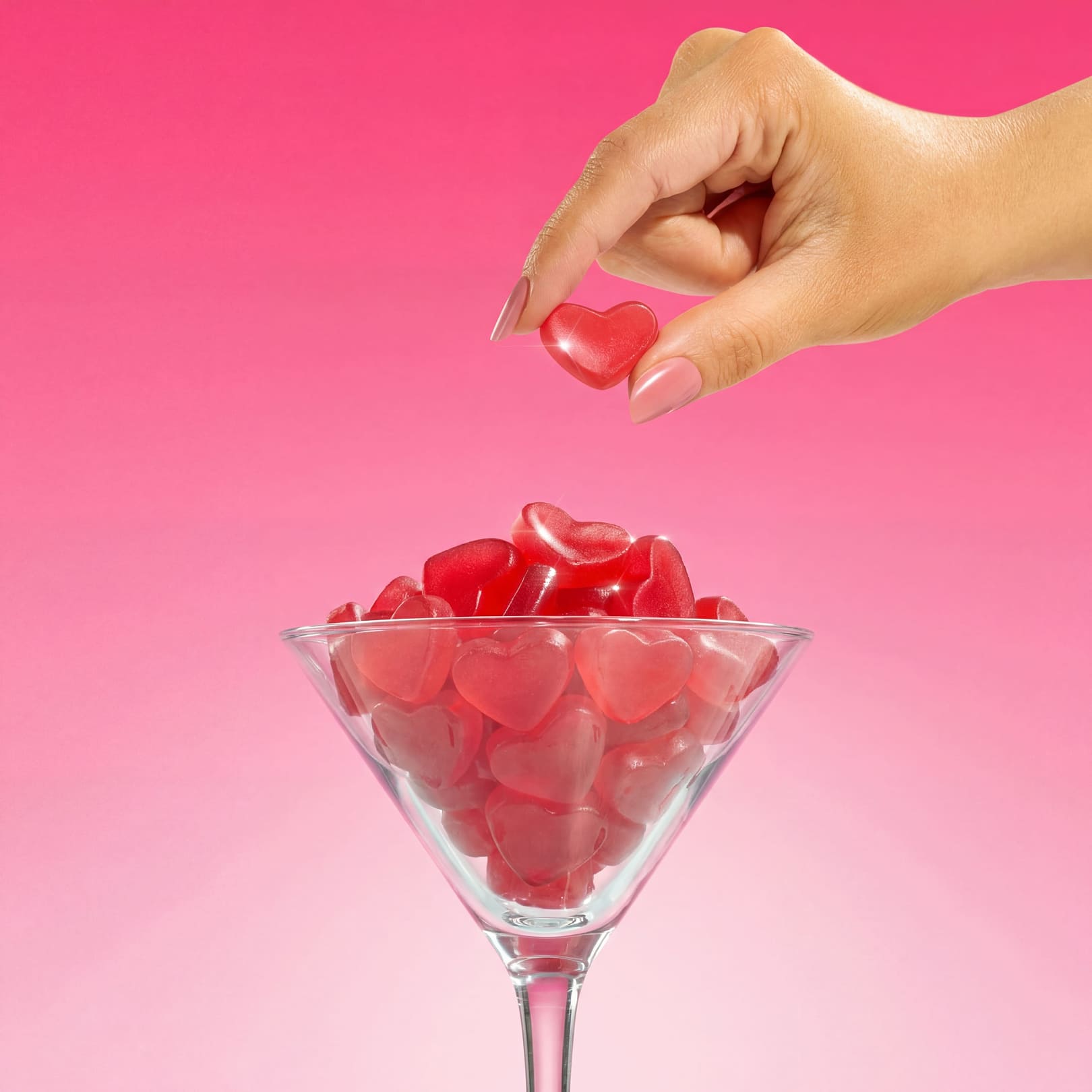 A hand holds a red, heart-shaped gummy over a martini glass filled with more heart-shaped gummies.