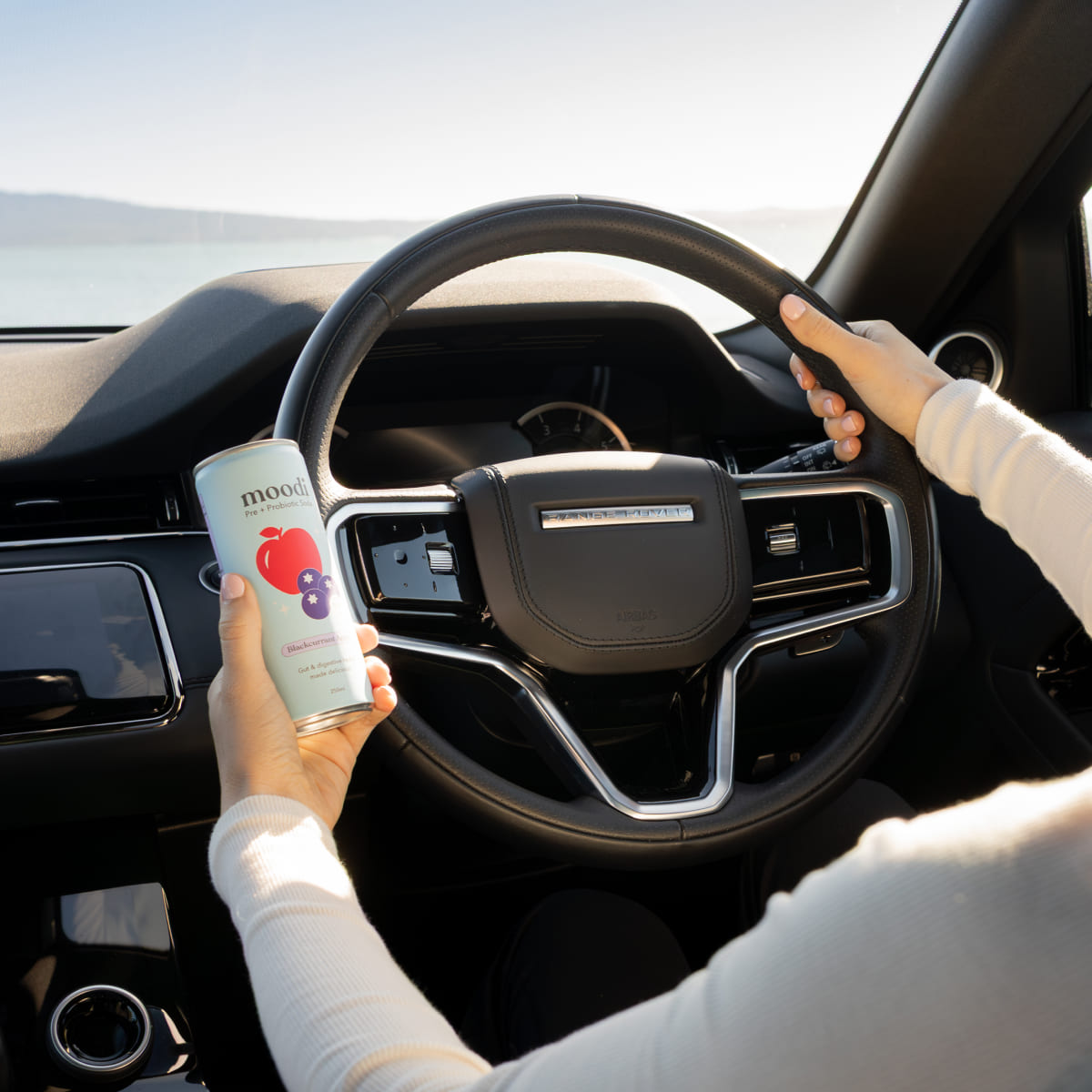Person holding a can while driving a car with a scenic view outside.