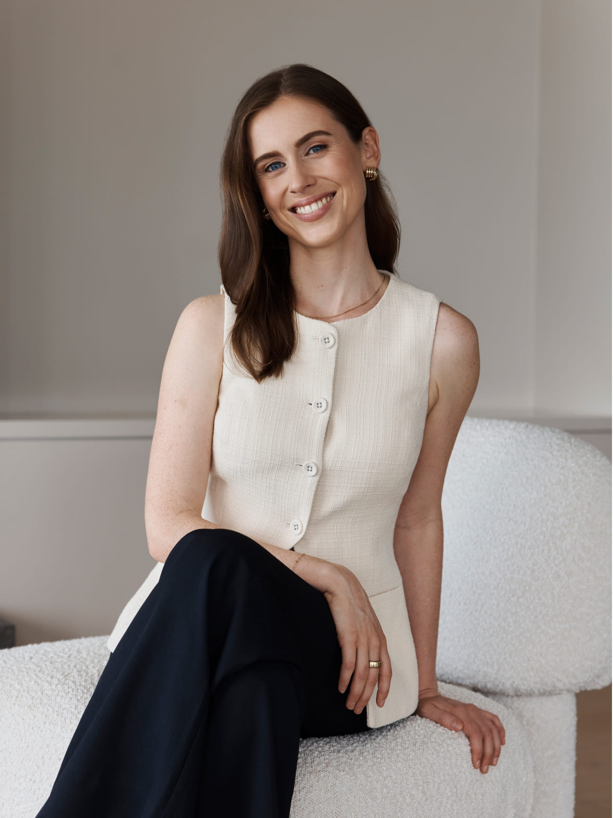 Smiling person seated on a white chair, wearing a sleeveless top and dark pants.