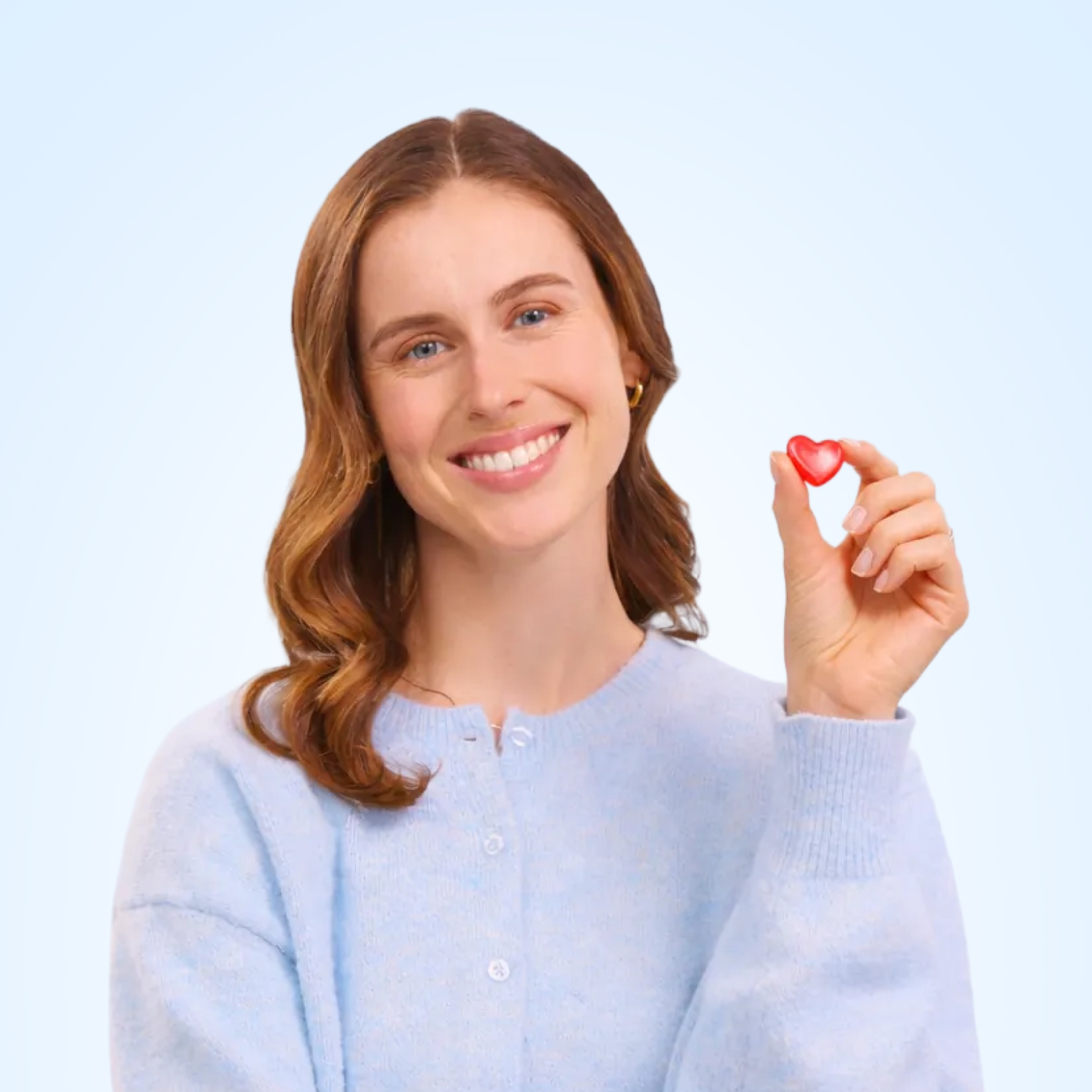 Person smiling, holding a small red heart-shaped object against a light blue background.