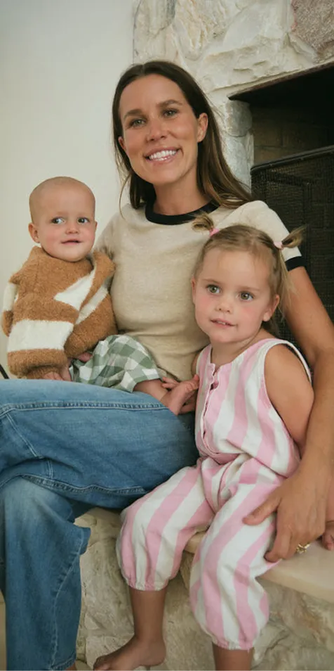 A smiling woman sits indoors with two young children, a baby and a girl, on her lap.