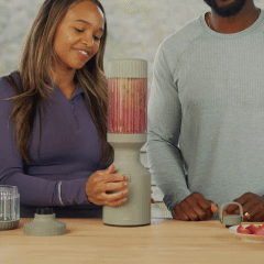 Two people using a blender with fruit on a kitchen counter.