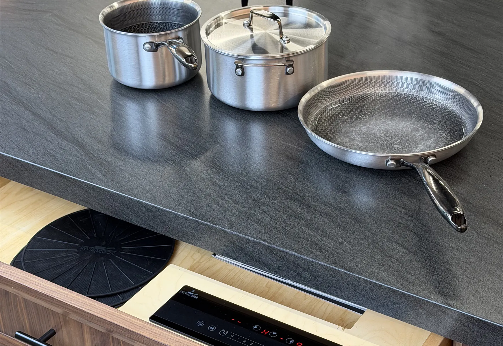 Stainless steel cookware sits on a dark countertop above an open drawer with an induction cooktop controller.