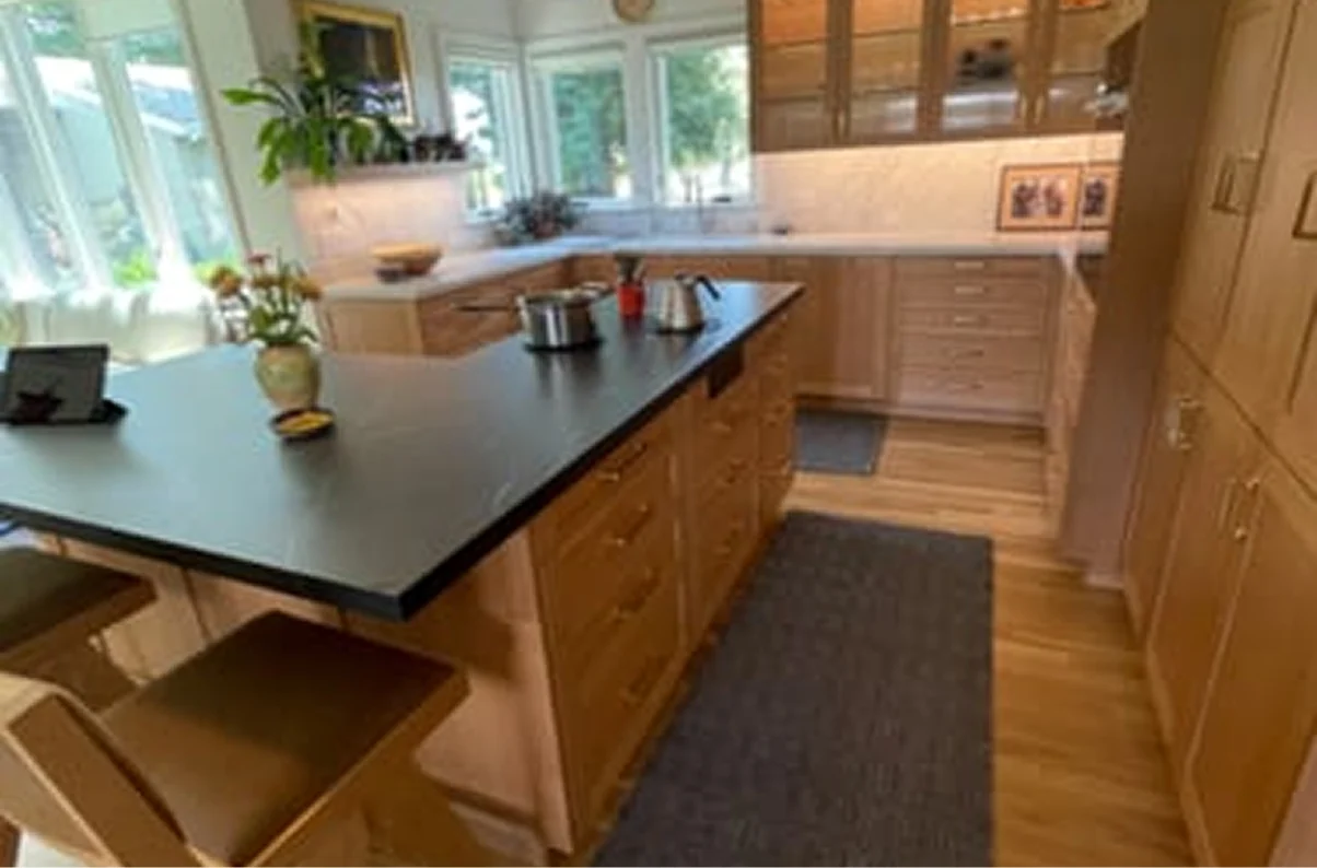 A spacious kitchen with light wood cabinetry, a large central island with a dark countertop, and large windows.