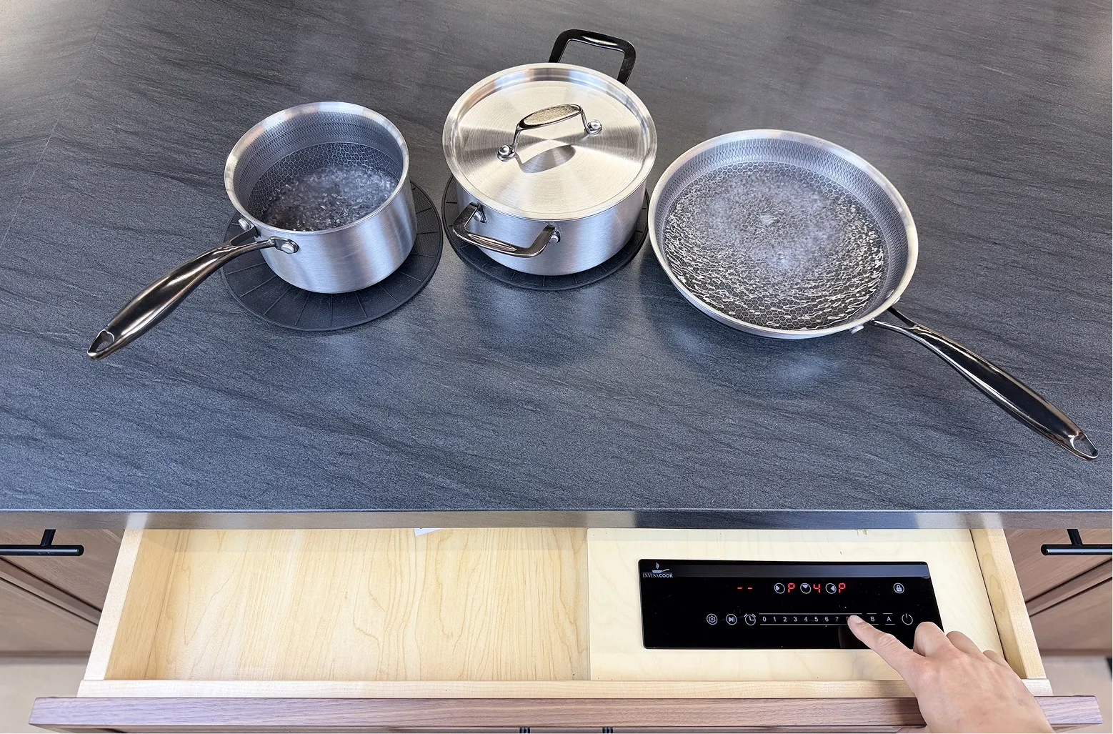Three pots of boiling water on a countertop with a hand adjusting controls inside an open drawer below.