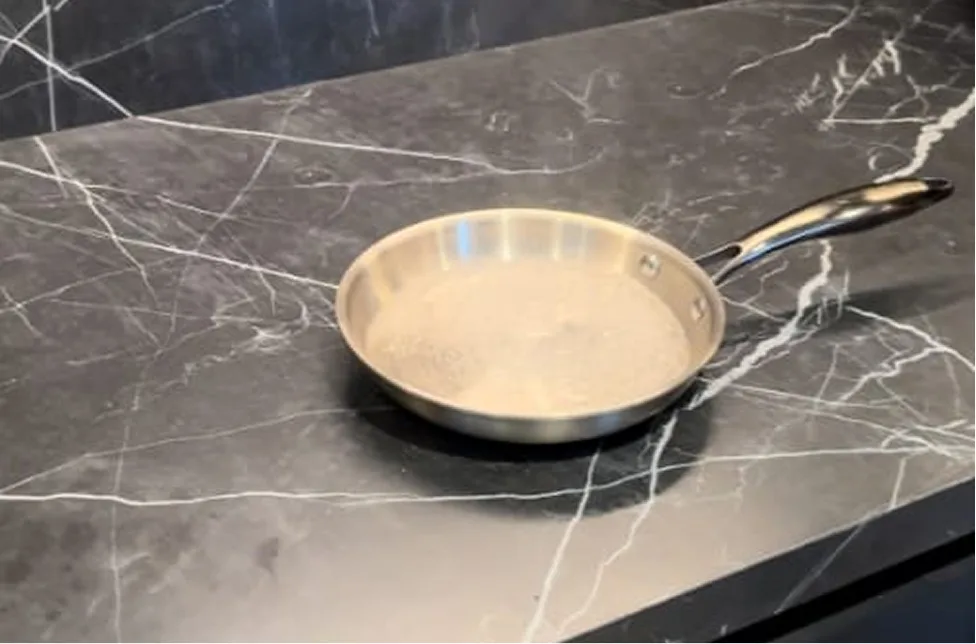 A stainless steel frying pan with a small amount of liquid sits on a black marble countertop.