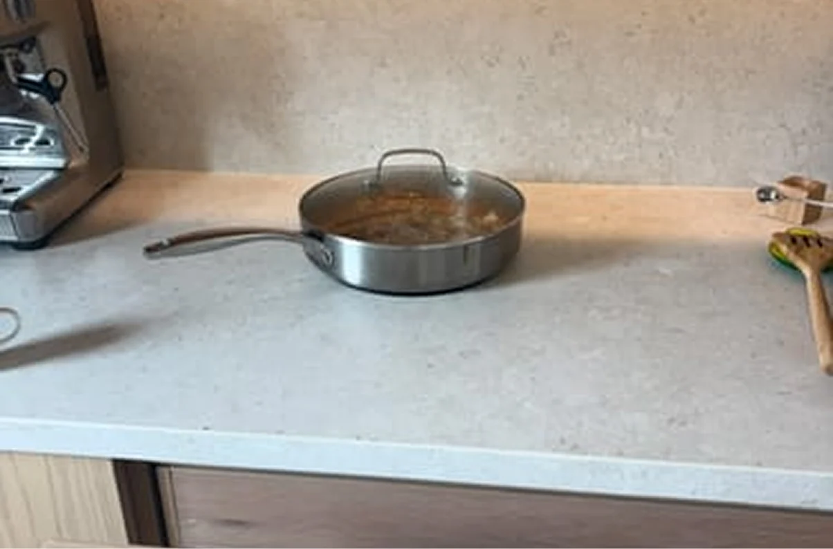 A stainless steel pan with a glass lid containing food sits on a light-colored kitchen countertop.