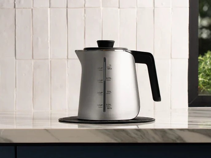 A silver electric kettle with a black handle sits on a marble countertop against a white tiled wall.