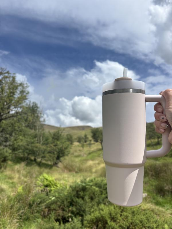 Hand holding a large beige tumbler outdoors with a cloudy sky and greenery in the background.