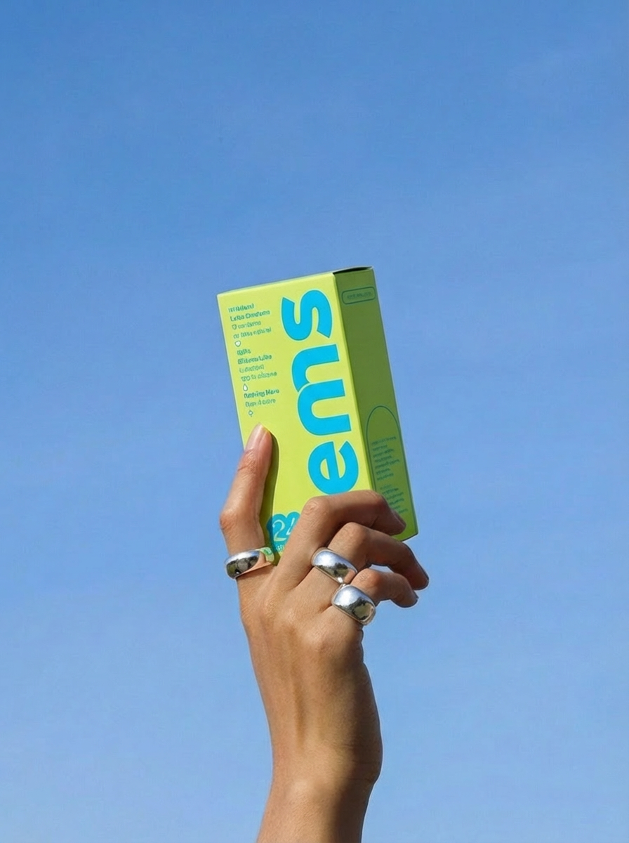 A hand wearing several silver rings holds a lime green box up against a clear blue sky.