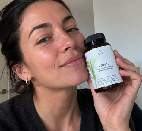 A woman with dark hair smiles while holding a bottle of 'Vitals' vitamins next to her cheek.