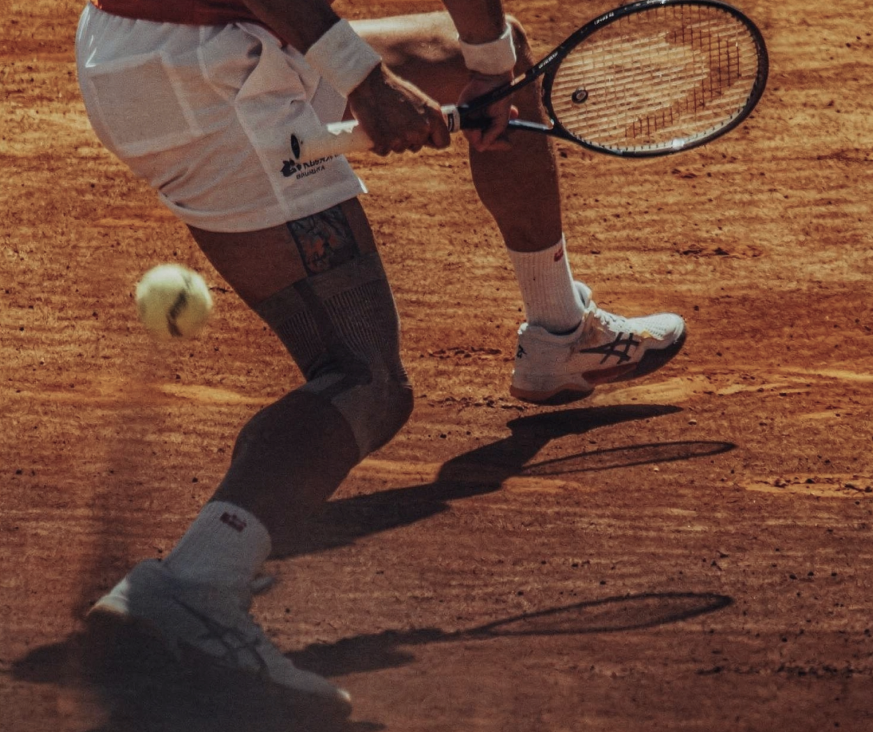 A close-up of a tennis player in white shorts hitting a ball on a clay court.
