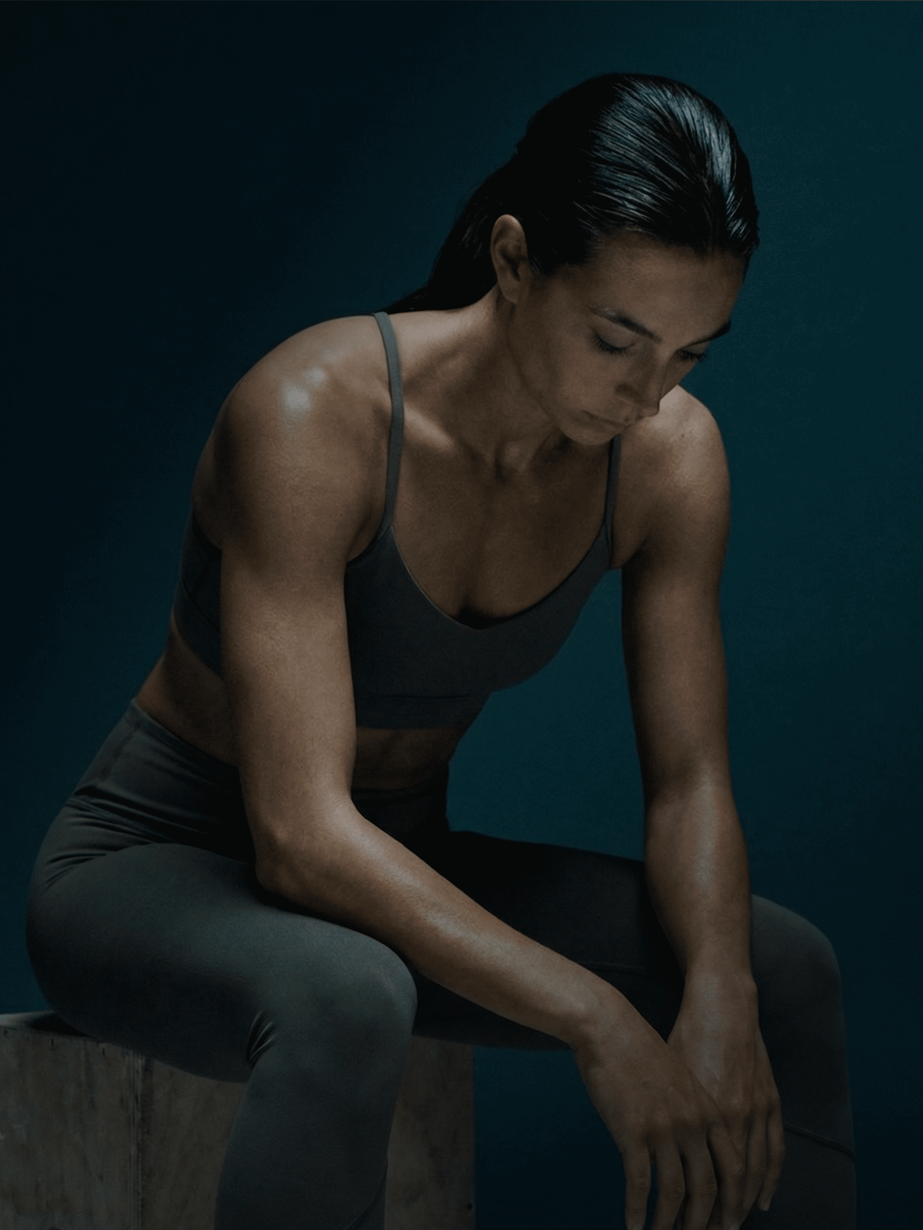 An athletic woman in workout clothes sits on a box with her head down, appearing to rest.