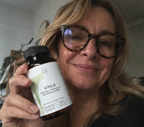 A smiling woman with glasses holds up a bottle of Vitals vitamins.