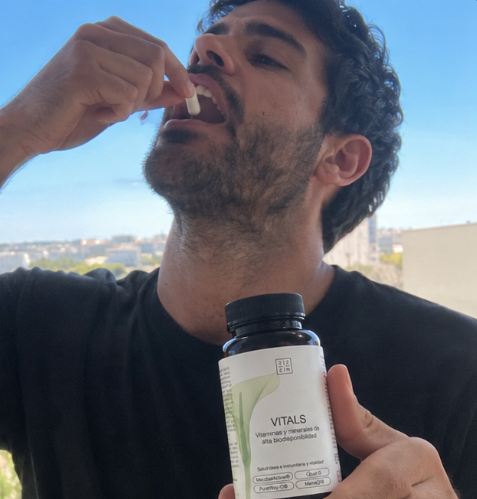 A man with a beard puts a white capsule in his mouth while holding a bottle of Vitals supplements.