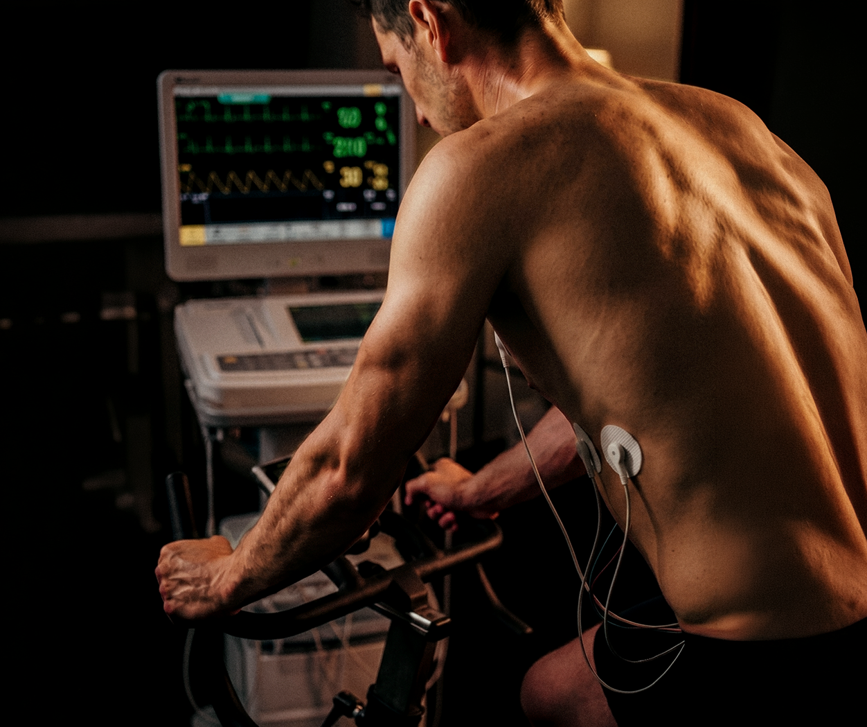 A man with electrodes on his back rides a stationary bike, facing a medical monitoring machine.