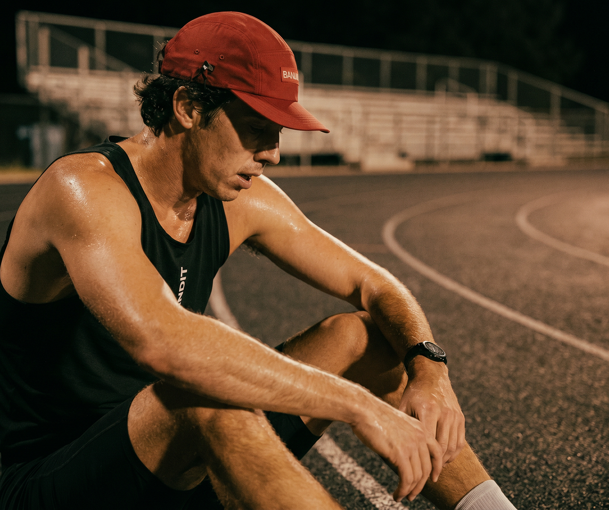 A sweaty man in a red cap sits on a running track at night, looking down.