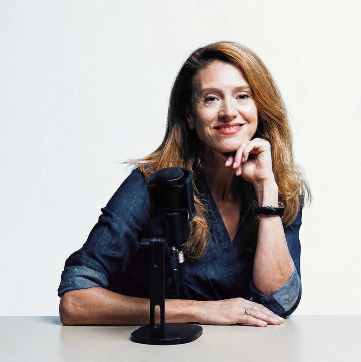 A smiling woman with long brown hair sits at a desk with a microphone, resting her chin on her hand.