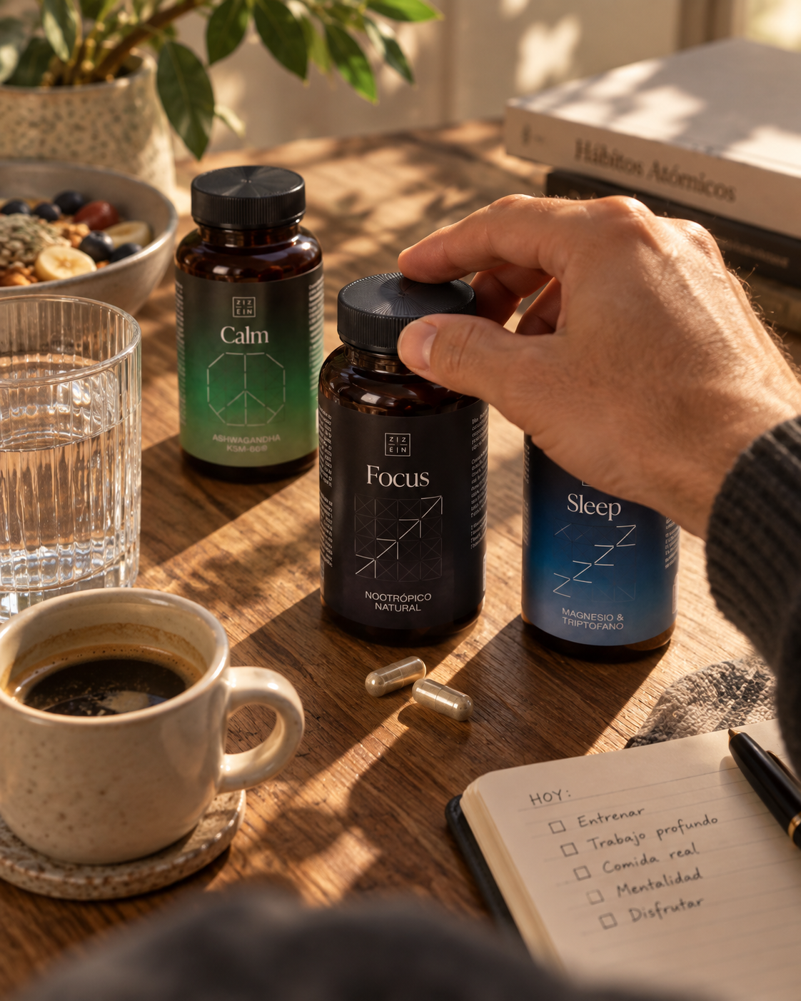 A hand reaches for supplement bottles on a wooden desk with coffee, water, and a notebook.