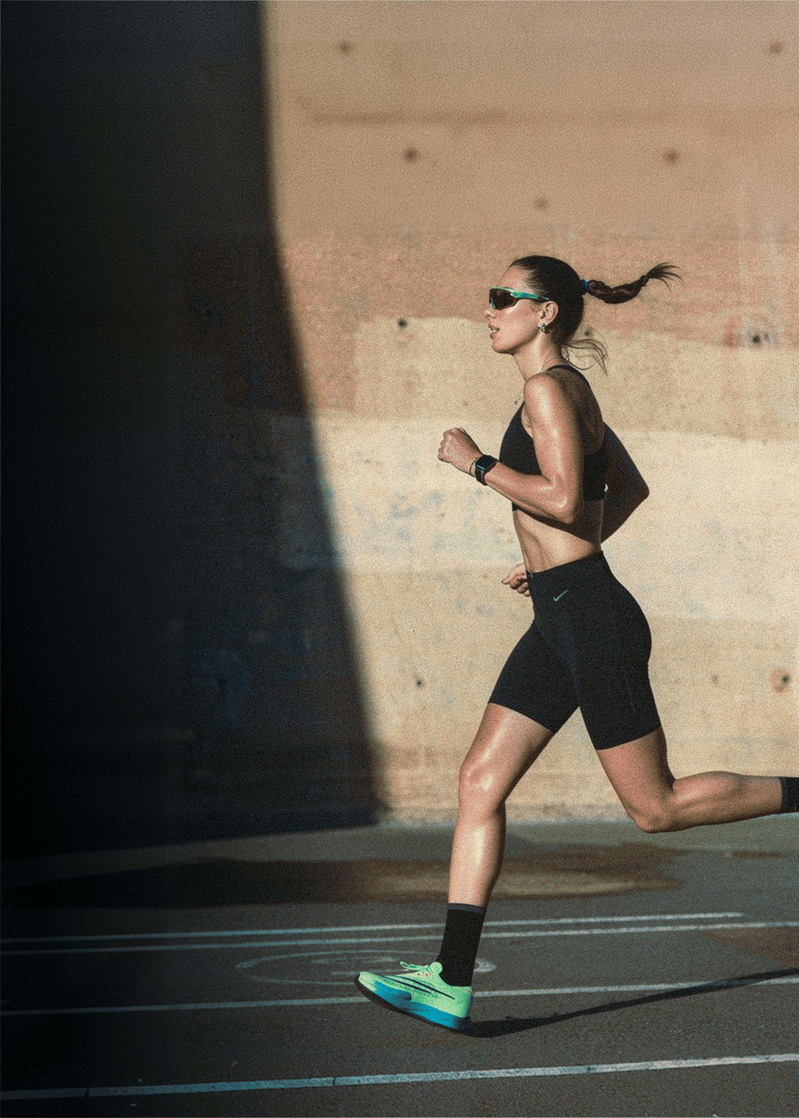 A woman in black athletic gear and sunglasses runs in profile past a concrete wall.