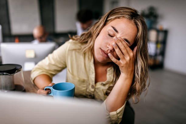 A tired-looking woman at her desk holds a blue mug and rubs her eyes with her hand.
