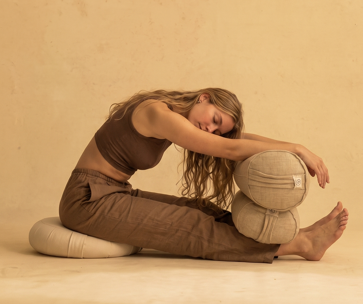A woman sits on a cushion, stretching forward with her arms and head resting on two stacked bolsters.