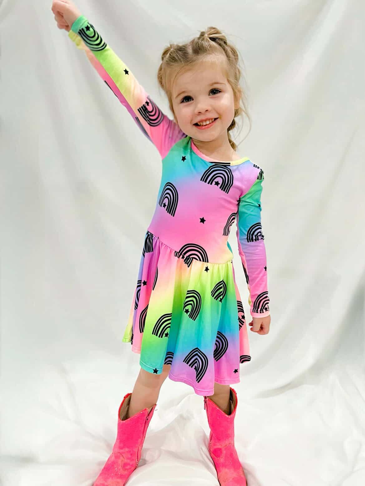 Child in colorful dress and pink boots, smiling and raising one arm.