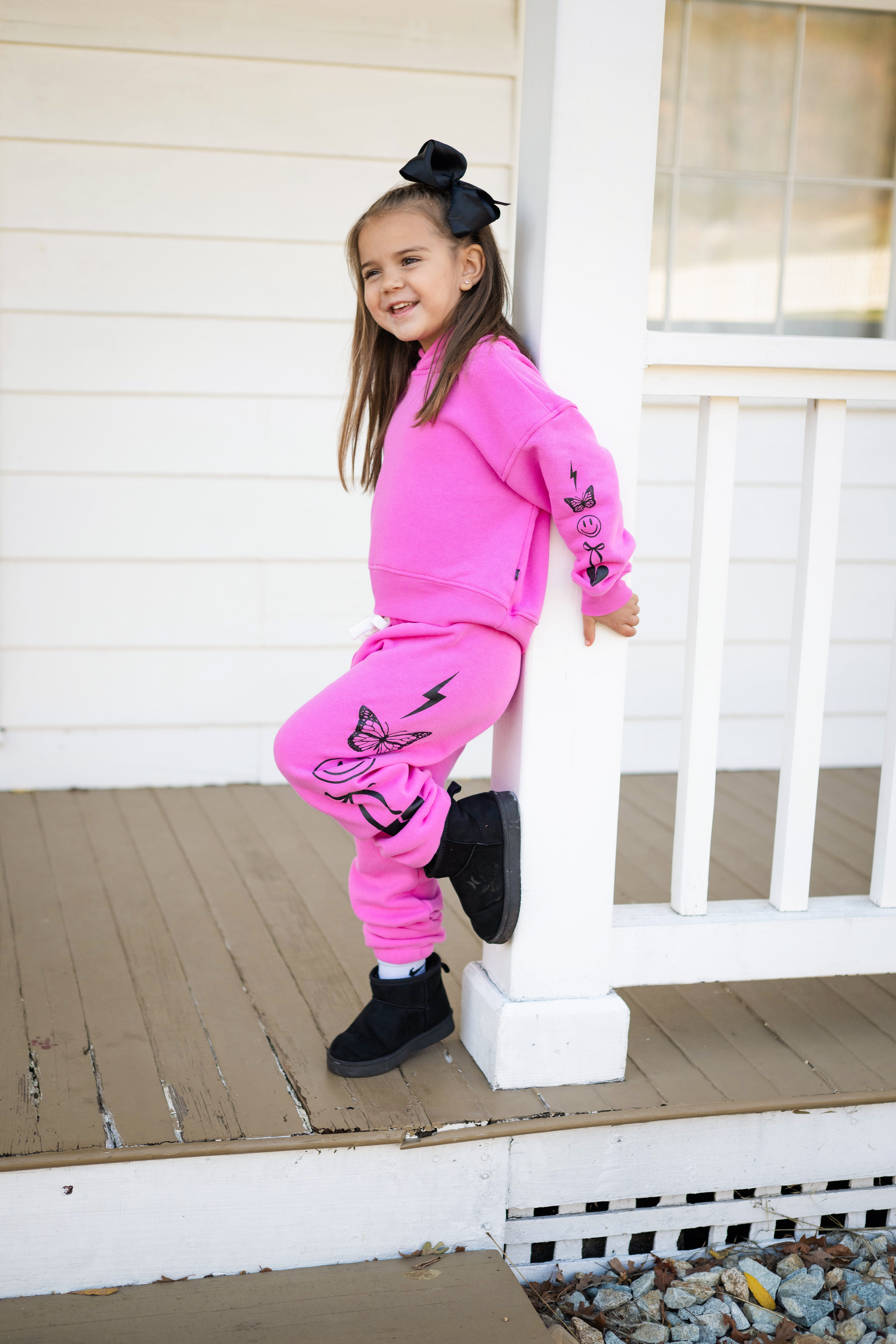 Child in pink outfit leaning on porch, smiling with hands in pockets.