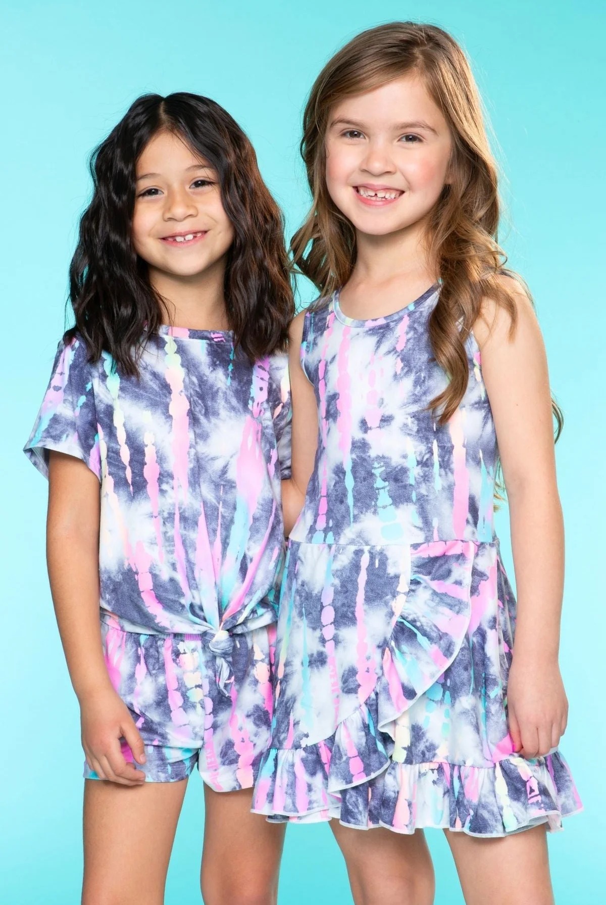 Two smiling girls in colorful tie-dye outfits against a blue background.