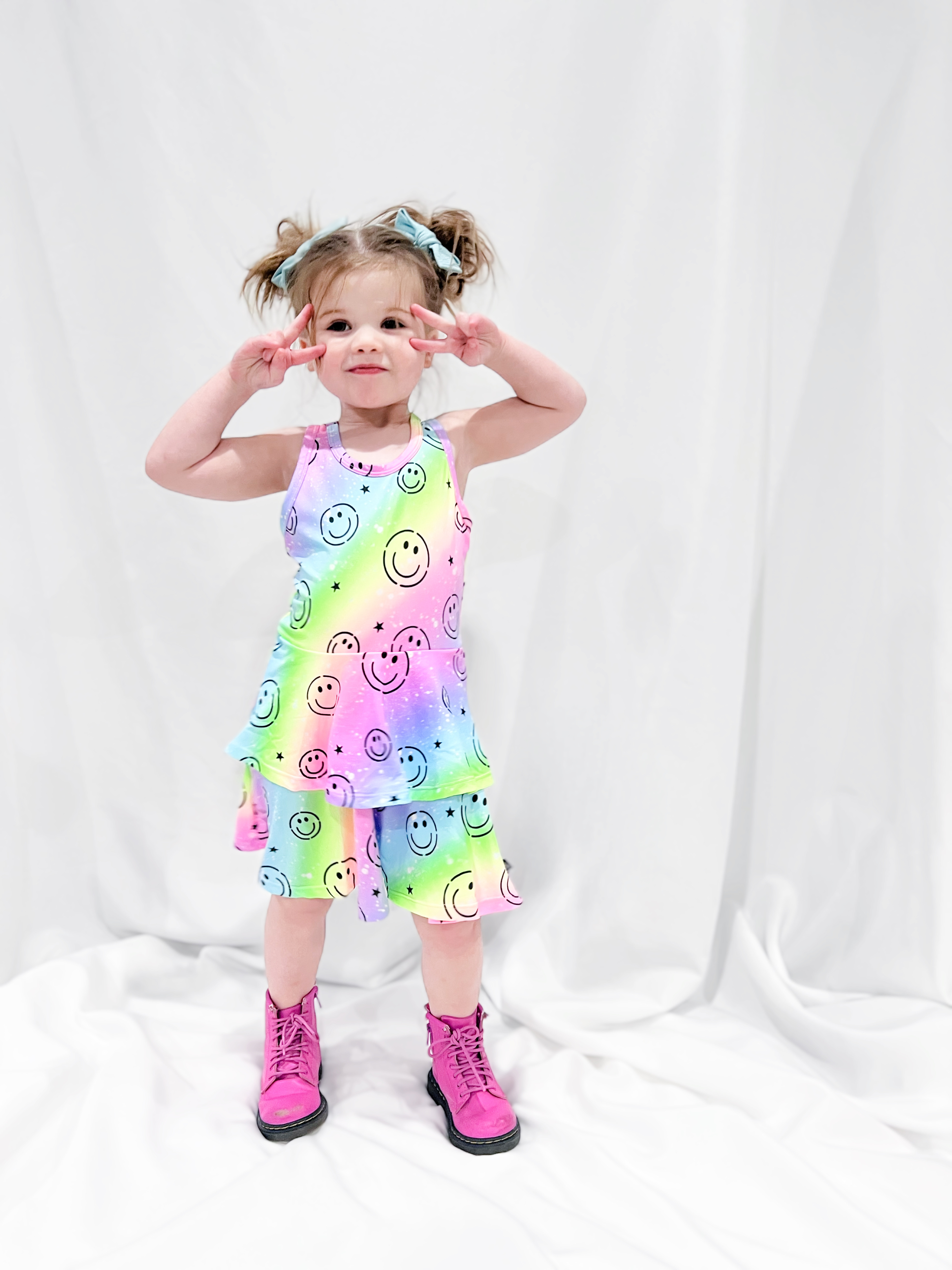 Child in rainbow dress with smiley faces, pink boots, and blue hair bows on white background.
