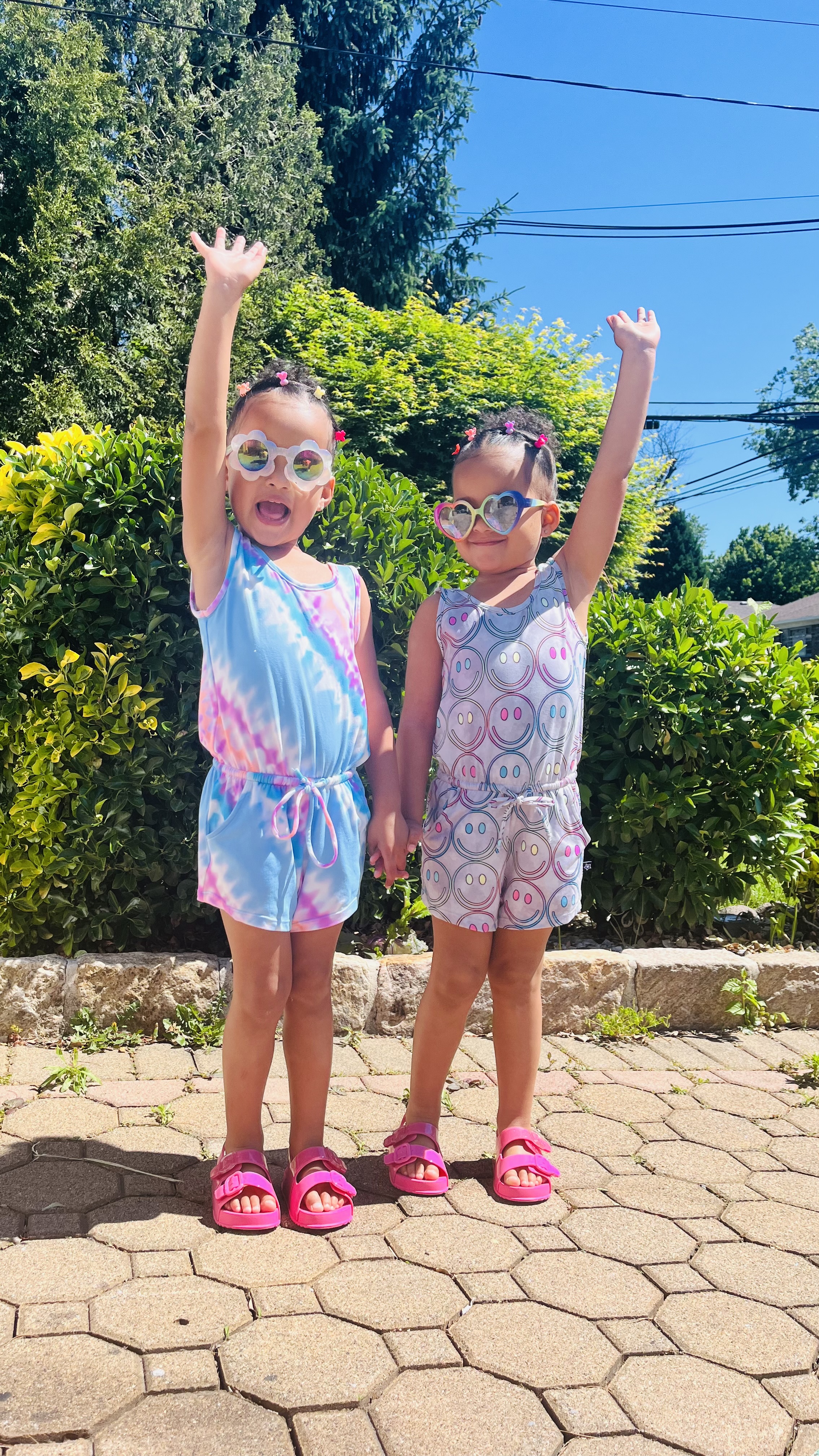 Two children in colorful outfits and sunglasses raising their hands outside on a sunny day.