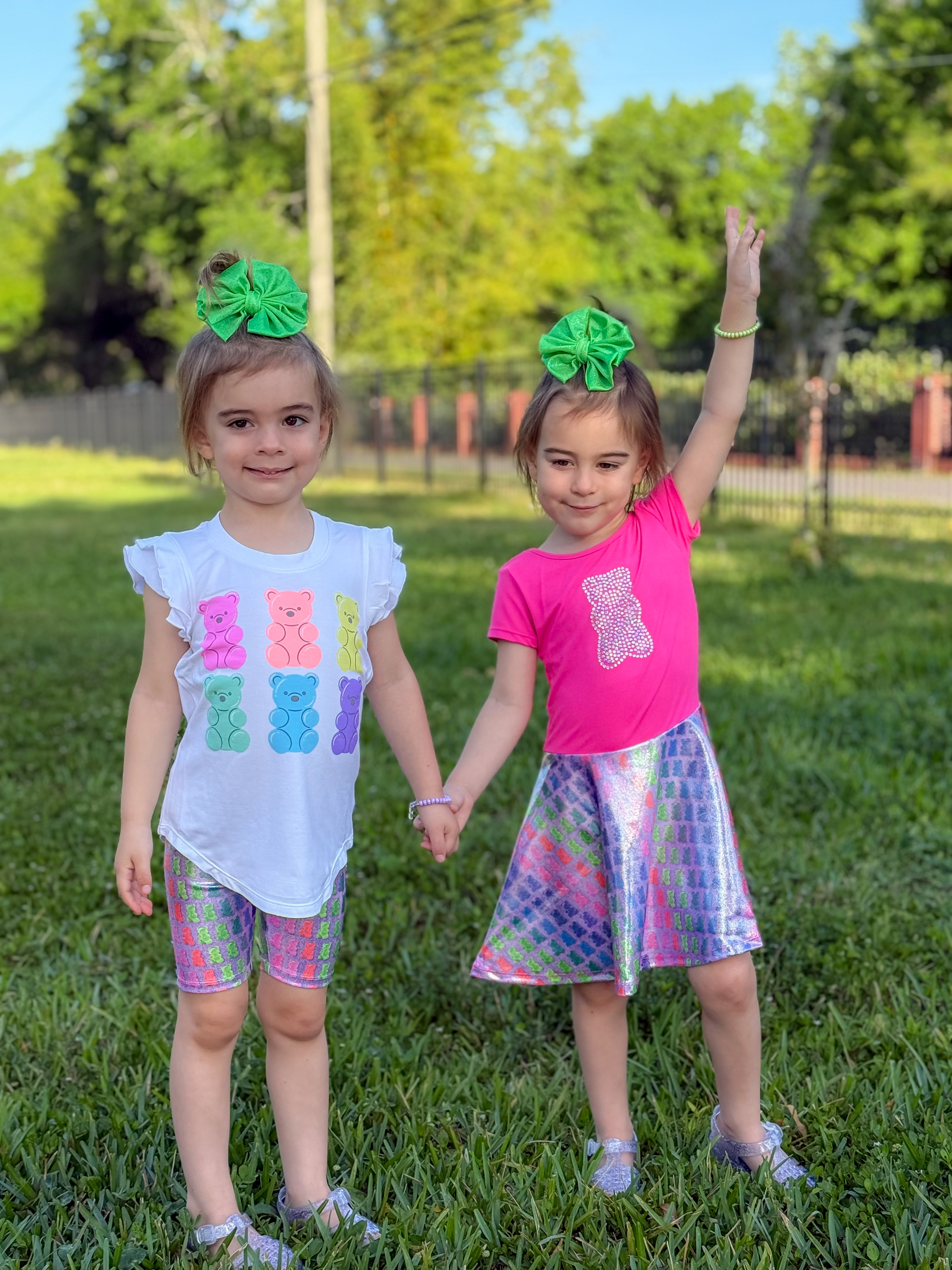 Two children holding hands in colorful outfits with green bows, standing on grass.