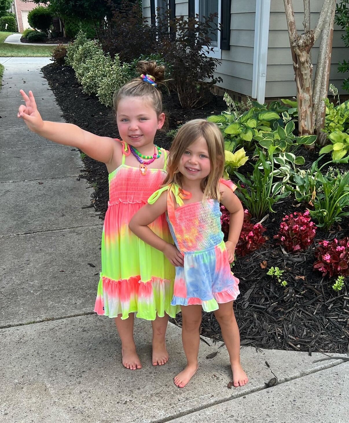 Two young girls in colorful dresses pose on a garden path.
