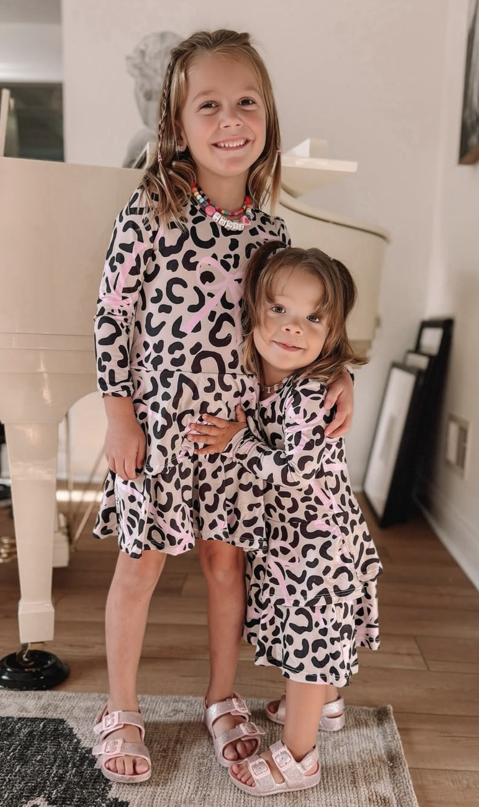 Two children in matching leopard print dresses standing by a piano.
