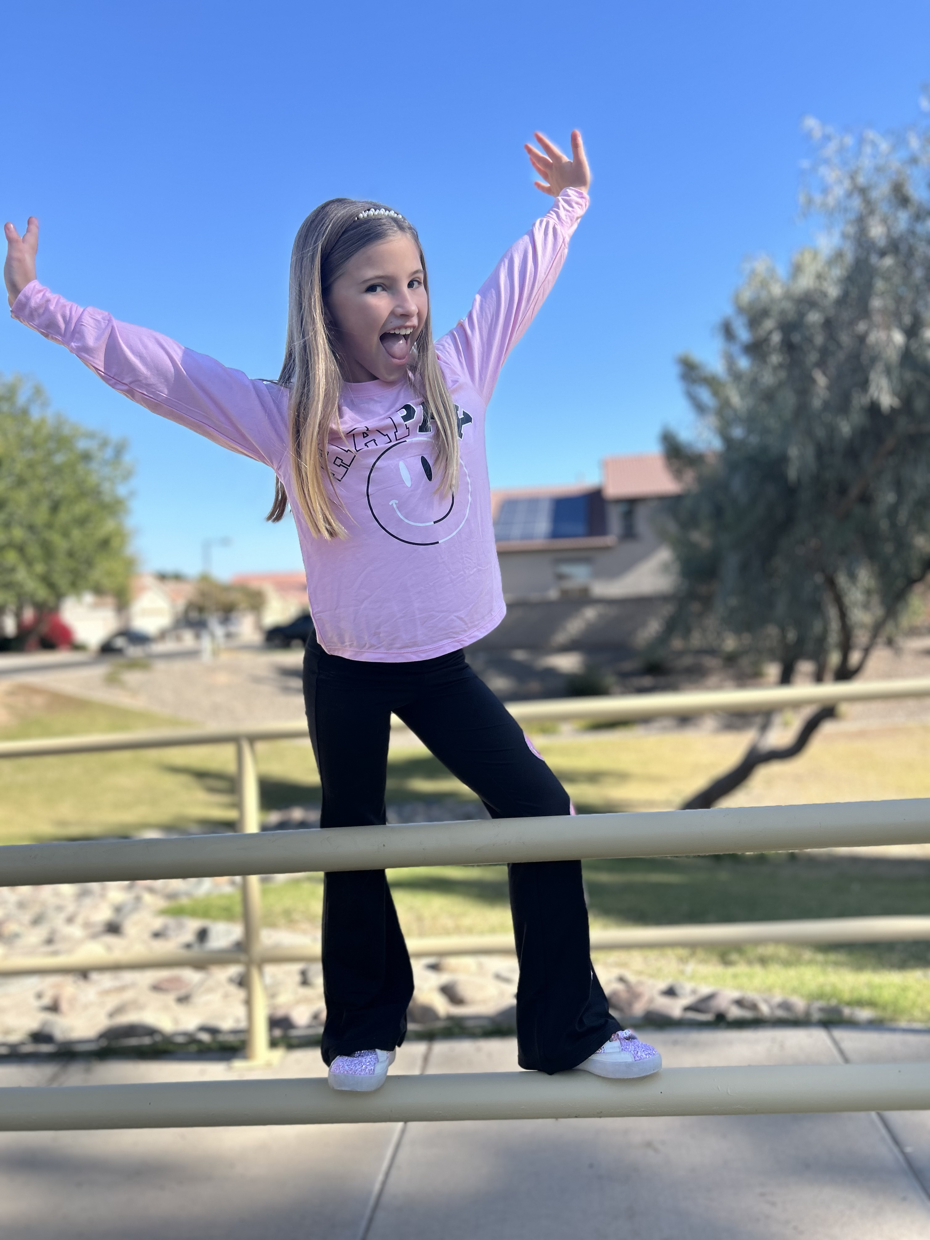 Child in pink shirt balancing on a yellow railing outdoors with trees in the background.