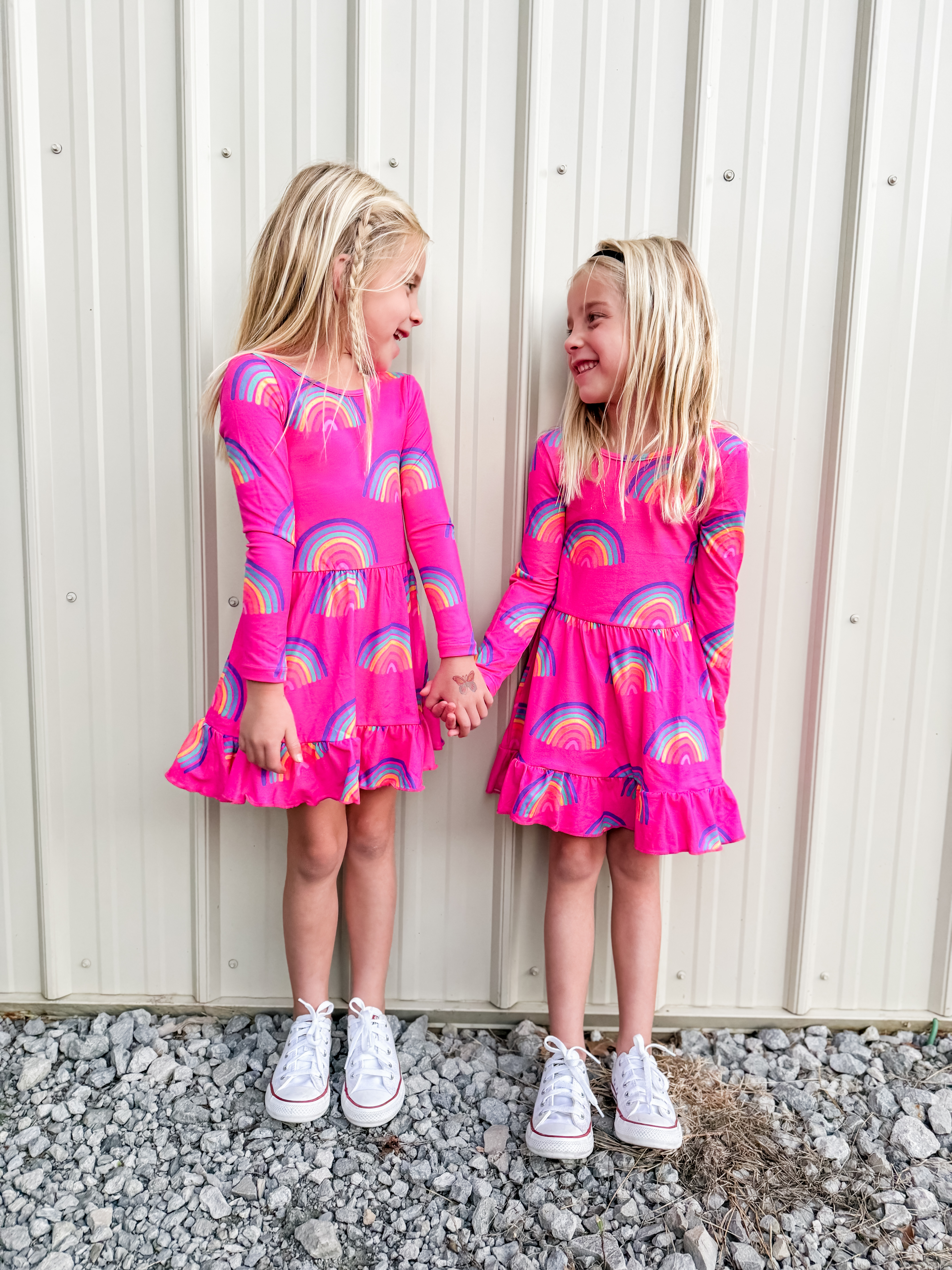 Two young children in pink rainbow dresses holding hands, standing against a metal wall.